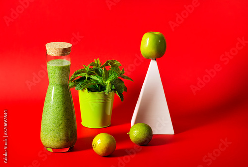 Fototapeta Naklejka Na Ścianę i Meble -  A beautiful bottle with a green smoothie stands on a red background, next to it stands a white geometric pyramid with a green apple, a bucket with mint leaves, and limes.