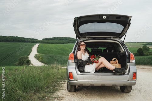 Young Stylish Woman in White Dress and Denim Jacket with Bouquet of Red Peonies Sits on the Trunk of Her SUV Car in the Green Field © Romvy