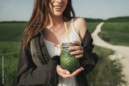 Young Stylish Girl in White Dress and Denim Jacket Holds Green Smoothie Fresh Drink in the Wheat Field