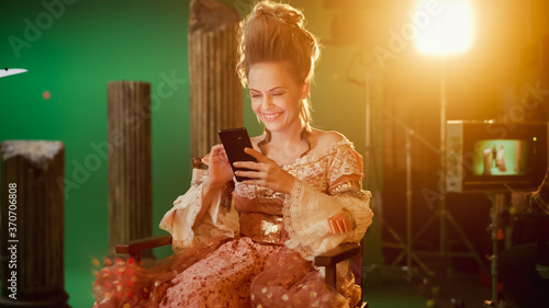 Fényképezés On Period Costume Drama Film Set: Beautiful Smiling Actress Wearing Renaissance Dress, Sitting on a Chair Using Smartphone with Green Screen in the Background