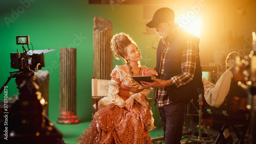 On Period Costume Drama Film Set: Beautiful Smiling Actress Wearing Renaissance Dress Sitting on a Chair Listens to Movie Director Explaining to Her Scene Context. High Budget Period Drama. photo