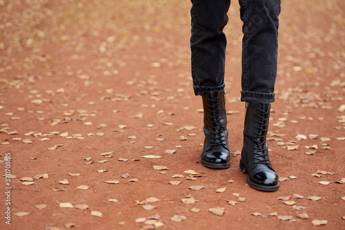 Woman s legs in fashionable autumn leather black boots and trendy black jeans. Trendy youth casual outfit for everyday. Modern street collection. Close-up. Autumn road with yellow leaves. Copy space.