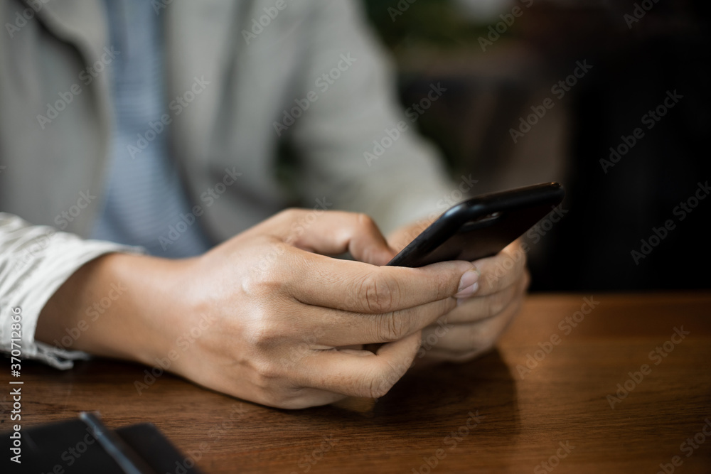 Fototapeta premium Close up of businessman's hands holding and typing on black cellphone for business communication in the cafe with a cup of coffe on wooden table. Business stock photo.