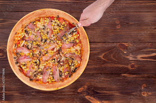 Man's hand cuts the pizza with chicken breast, corn, bacon and mushrooms, with a pizza cutter on round wood plate which is on wooden rustic table, top view and copy space