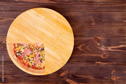 Quarter of pizza with chicken breast, corn, bacon and mushrooms, on a round wood plate which is on wooden rustic background, top view and copy space
