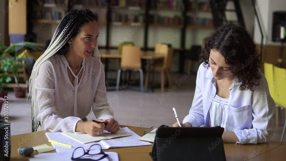 Portrait of two attractive women having a conversation in a library with shelves of books on background. Sitting at the wooden table with tablet and diverse papers. Dreadlocks woman explaining smth Portrait of two attractive women having a conversation in a library with shelves of books on background. Sitting at the wooden table with tablet and diverse papers. Dreadlocks woman explaining smth