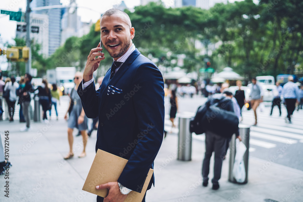 Cheerful businessman in formalwear talking on phone walking on street getting to work, successful financial manager excited with good news during mobile conversation outdoors in megalopolis.