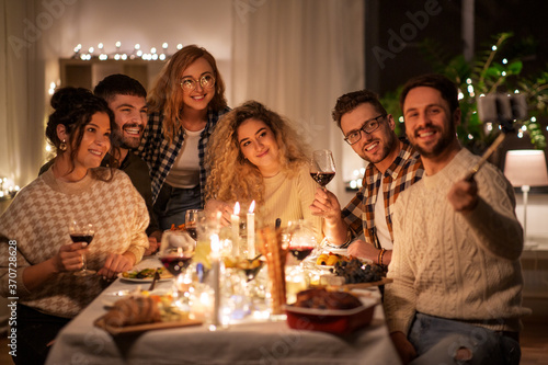 holidays  celebration and people concept - happy friends taking with smartphone on selfie stick at home christmas dinner party