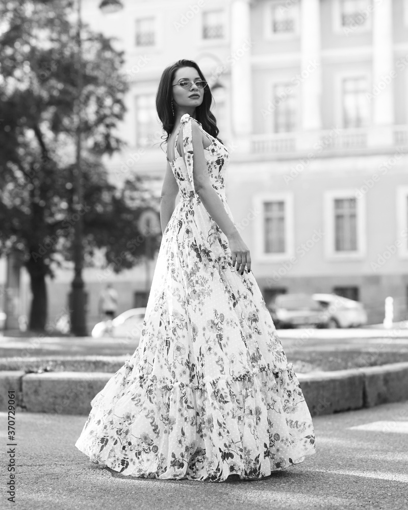 Young beautiful woman wearing white dress with floral prink and walking at city street between columns on a sunny day. Female model with long wavy brunette hair Young beautiful woman wearing white dress with floral prink and walking at city street between columns on a sunny day. Female model with long wavy brunette hair