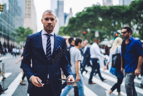 Portrait of serious male entrepreneur standing on crosswalk in new york city looking at camera, confident businessman dressed in elegant suit using modern technology while walking in office .