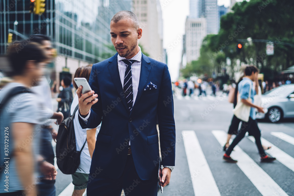 Thoughtful male owner reading information from websites on smartphone concentrated on news while pedestrians passing by, confident manager sending messages via mobile while standing Thoughtful male owner reading information from websites on smartphone concentrated on news while pedestrians passing by, confident manager sending messages via mobile while standing
