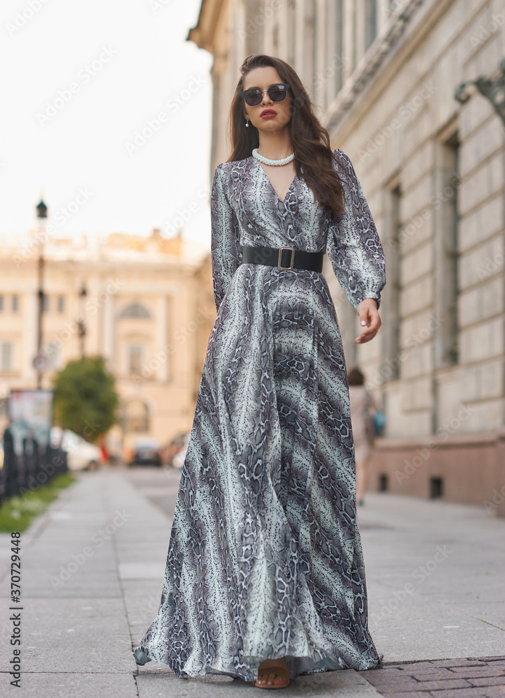 Naklejka premium Pretty female model in gray dress with stripes and sunglasses standing and posing at city street on a summer day. Full length fashion oudoor portrait