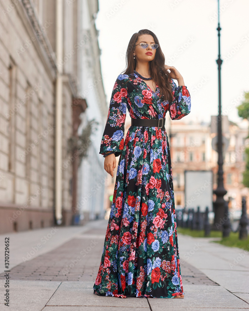 Young elegant lady in red and black floral design dress. Full length woman portrait. Beautiful girl standing and posing at city street. Fashionable female model with wavy hair
