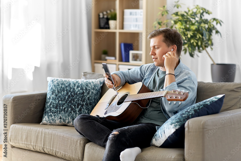 leisure, music and people concept - young man or musician with guitar, earphones and smartphone sitting on sofa at home