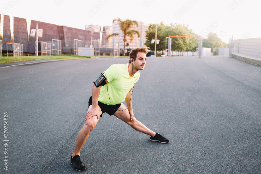 Full length portrait of young sportsman in bright sports clothing makes exercises for legs before start morning run in an urban setting, handsome athlete playing sports on the road in summer morning