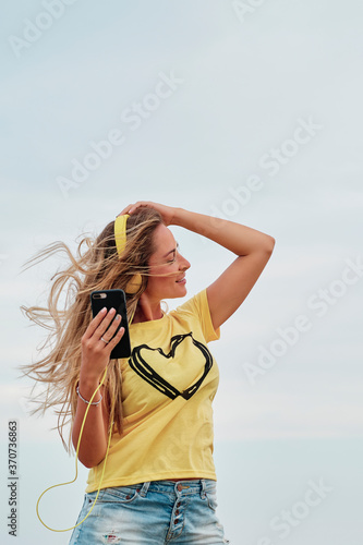 young blonde woman using yellow headphones in front of the beach on a summer cloudy day