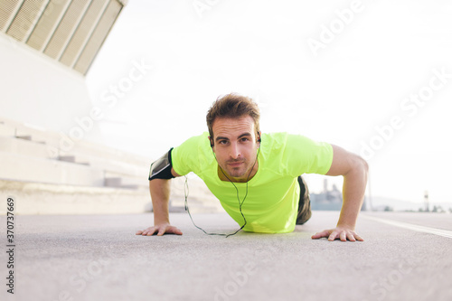 Portrait of strong build sports man doing push-ups on asphalt city road and listen to music with headphones, young male jogger warm up before start his workout training outside and look to the camera