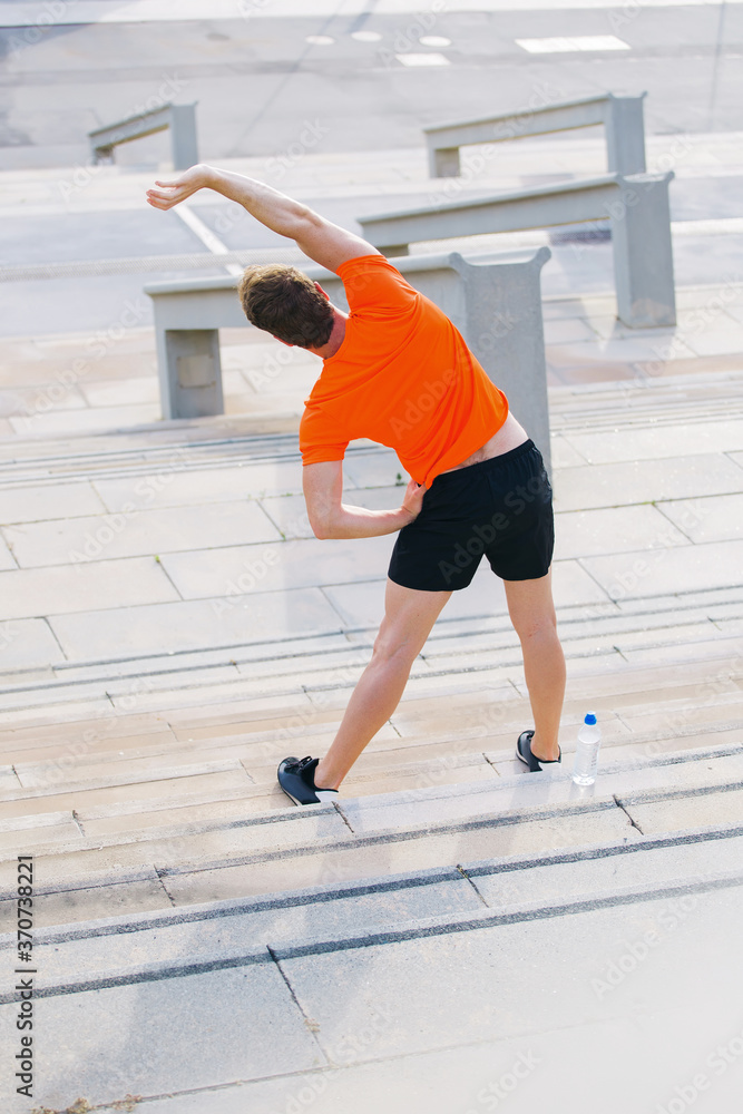 Back view of fit men stretches muscles before morning jog outdoors in summer day, young sportsmen dressed in sportswear workout in the fresh air, male runner engaged in physical exercises outdoors Back view of fit men stretches muscles before morning jog outdoors in summer day, young sportsmen dressed in sportswear workout in the fresh air, male runner engaged in physical exercises outdoors