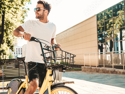 Portrait of handsome smiling stylish hipster lambersexual model.Man dressed in white T-shirt. Fashion male riding a bike on the street background in sunglasses © halayalex