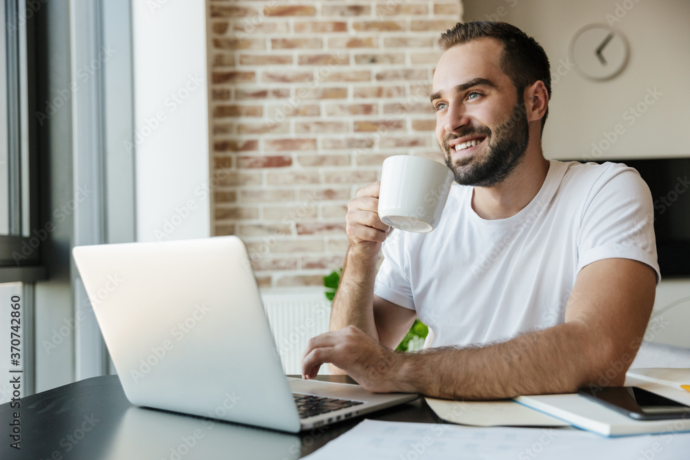 Image of man drinking coffee and using laptop while sitting at table Image of man drinking coffee and using laptop while sitting at table