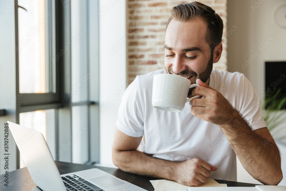 Image of man drinking coffee and using laptop while sitting at table Image of man drinking coffee and using laptop while sitting at table