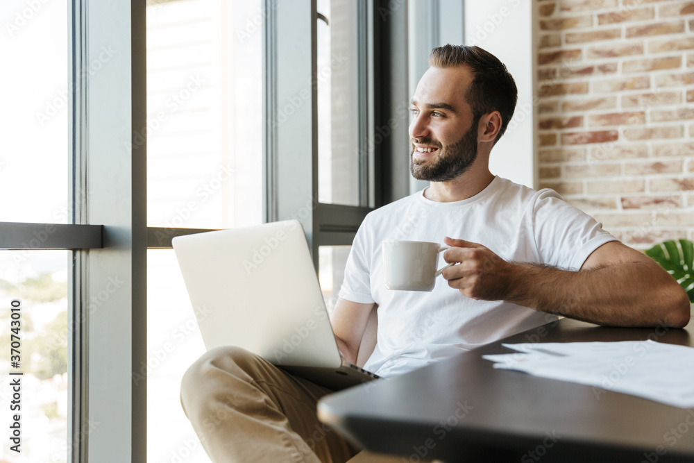 Image of man drinking coffee and using laptop while sitting at table Image of man drinking coffee and using laptop while sitting at table