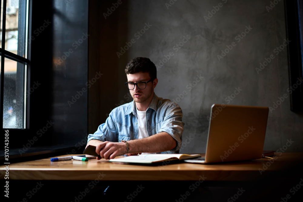 Professional designer dressed in denim shirt holding telephone in hand while working on new graphic project during working process sitting in own studio with laptop computer connecting Professional designer dressed in denim shirt holding telephone in hand while working on new graphic project during working process sitting in own studio with laptop computer connecting