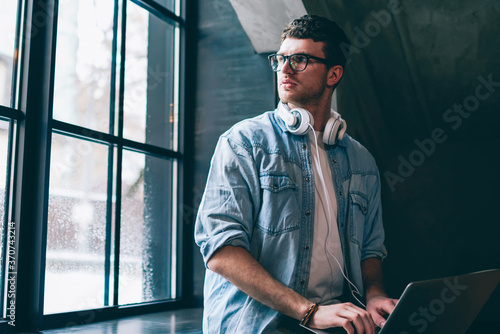 Young contemplative male freelancer pondering on program code holding laptop during free time at coworking space, thoughtful copywriter in casual apparel looking away using modern technology indoors