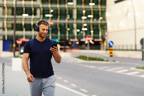 Young sport man walking in the city and listens to music via smartphone through wireless headphones