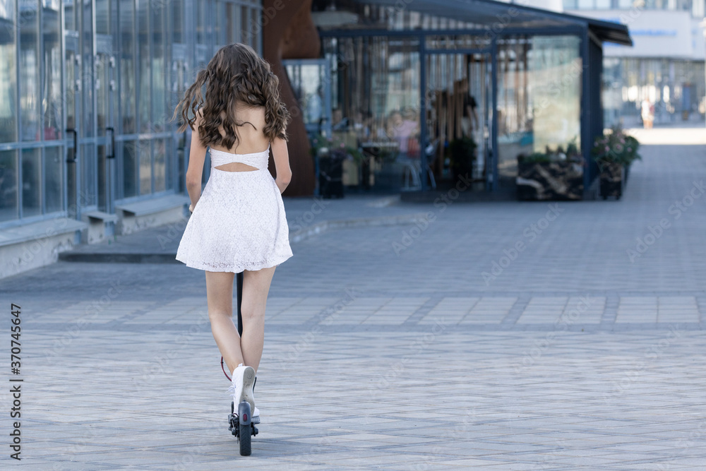 Naklejka premium A girl in a dress who rides an electric scooter in the city