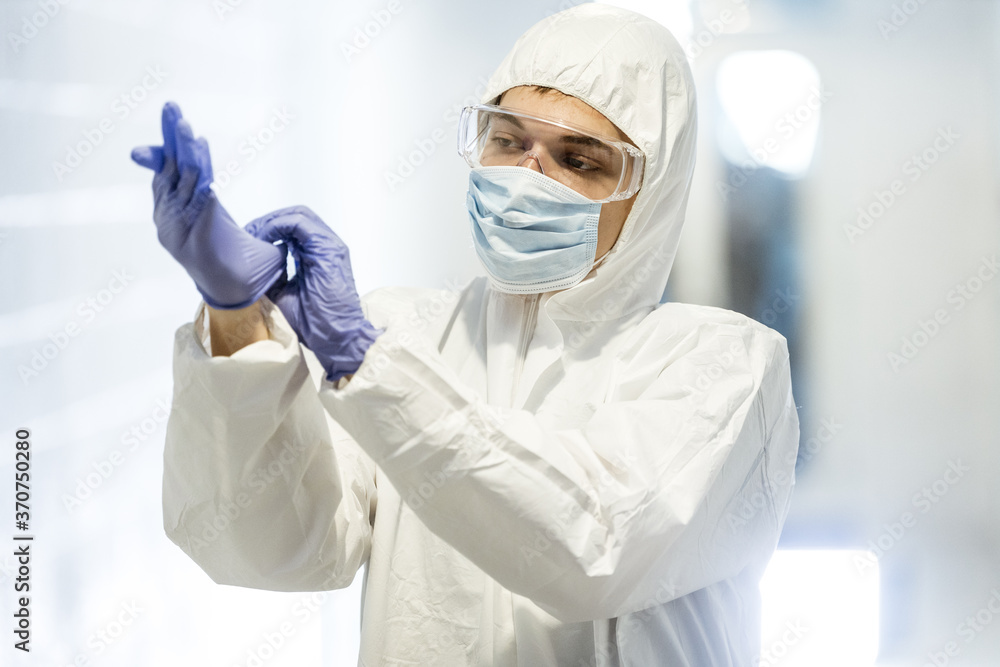 Corona virus concept. Male scientist doctor in mask, glasses and protective suit putting on latex gloves and getting ready for COVID-19 pandemic outbreak quarantine. Man in biochemistry laboratory Corona virus concept. Male scientist doctor in mask, glasses and protective suit putting on latex gloves and getting ready for COVID-19 pandemic outbreak quarantine. Man in biochemistry laboratory