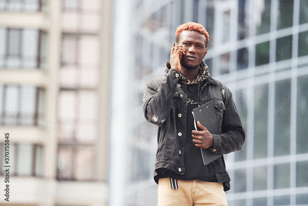 Talking by phone. Young african american man in black jacket outdoors in the city standing against modern business building Talking by phone. Young african american man in black jacket outdoors in the city standing against modern business building