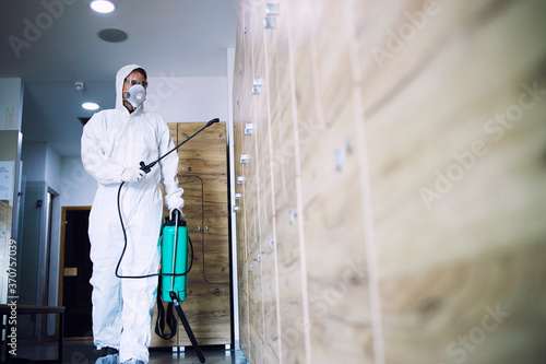 Public areas disinfection and healthcare. Man in white protection suit disinfecting and sanitizing lockers in gym's dressing room to stop spreading highly contagious coronavirus or COVID-19. © littlewolf1989