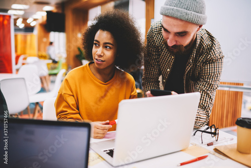 Two creative international male and female students in casual apparel thinking on productive developing own startup project teamworking at modern laptop in office during studying brainstorming