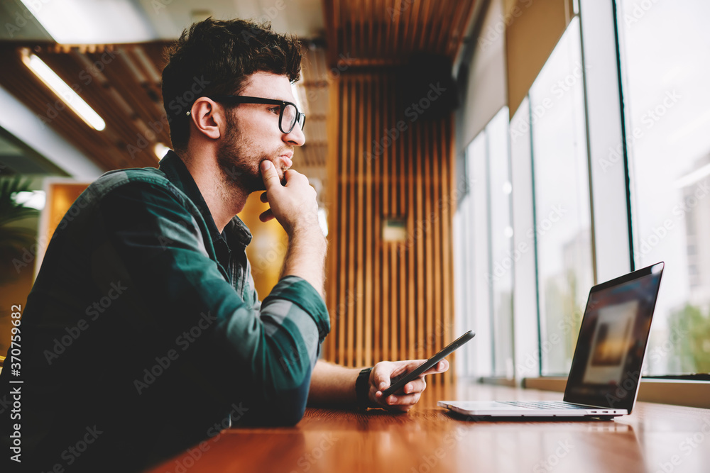 Young professional IT developer wearing eyeglasses for vision correction pondering about creative ideas for startup.Bearded hipster guy planning schedule while installing application on laptop