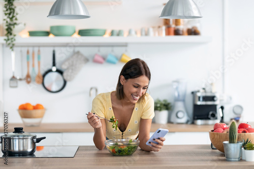 Beautiful young woman using her mobile phone while eating a salad in the kitchen at home.