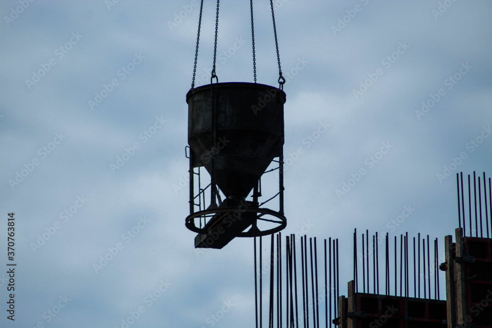 Concrete mixer at a construction site raised on a crane against the background of iron fittings and overcast sky Concrete mixer at a construction site raised on a crane against the background of iron fittings and overcast sky