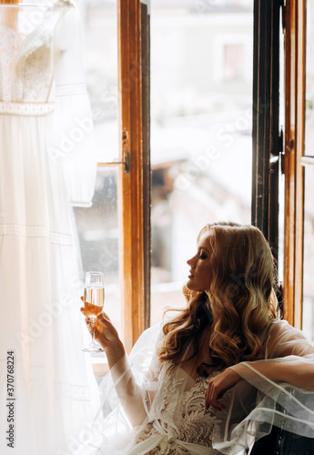 The elegant bride is sitting in front of the window holding a glass of champagne in her hands. Morning of the bride