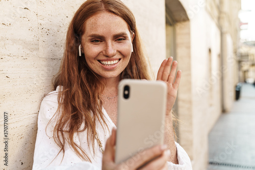 Image of joyful ginger woman waving hand and using cellphone