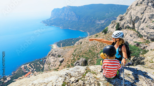 Child, young mother sit on mount top. Look at amazing sea landscape. Family travel adventure, hiking activity. Via ferrata tour with kids, exploring nature on summer vacation. Weekend day walking tour photo
