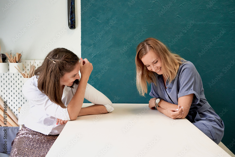 Two beautiful women laugh. Female young businesswoman sit at table in workplace and talk, have fun and gossip Two beautiful women laugh. Female young businesswoman sit at table in workplace and talk, have fun and gossip