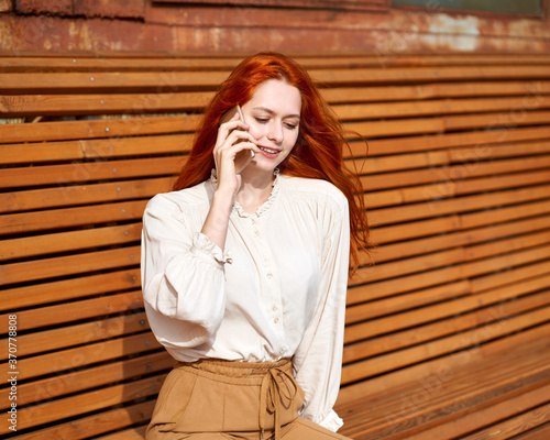 Red-haired woman talking on cell phone. Beautiful stylish fashion model sitting on bright orange bench in sun on summer day