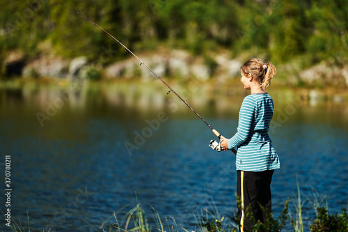 A young girl fishes at a beatiful lake in the deep and wild forest of Norway. Alone in the relaxing nature.