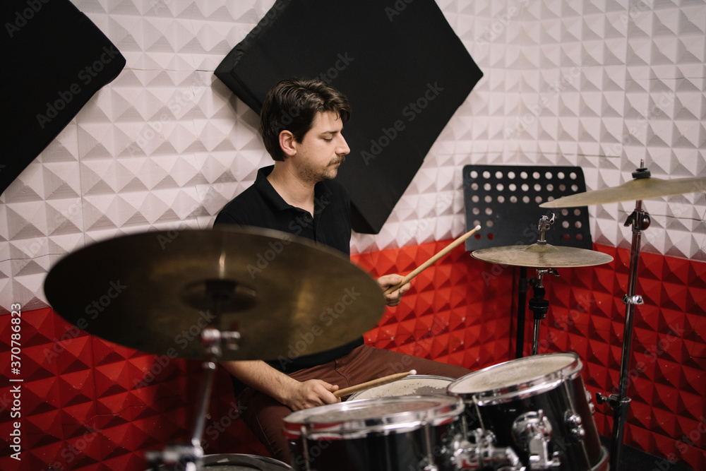 Fototapeta premium Young male drummer playing drums and cymbals. Handsome man with moustache playing with drumsticks while sitting at drums in studio.