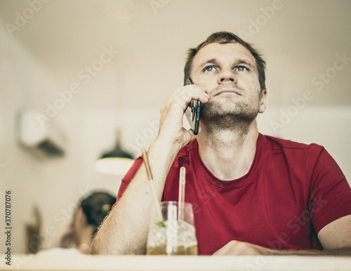 Restrained smile serious young man answer smart phone in red shirt in cafe drinking ice tea, waiting somebody or coworking. Real people expression, life problems solving or communication, relationship