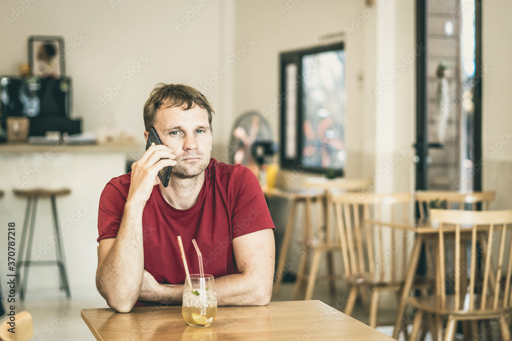 Serious sad young man answering smart phone in red shirt in cafe drinking ice tea, waiting somebody or coworking. Real people expression, life problems solving or communication, relationship Serious sad young man answering smart phone in red shirt in cafe drinking ice tea, waiting somebody or coworking. Real people expression, life problems solving or communication, relationship