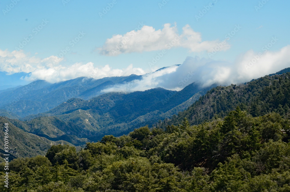 Beauful mountain landscape, white puffy clouds hover over part of mouintain range, looks like mountain has a blanket of clouds on, green slopes, blue sky and inspiring.
