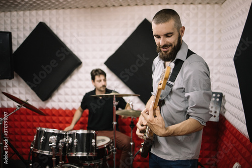 Music band playing instruments at soundproof studio. Two band members playing electric guitar and drummer set and recording.