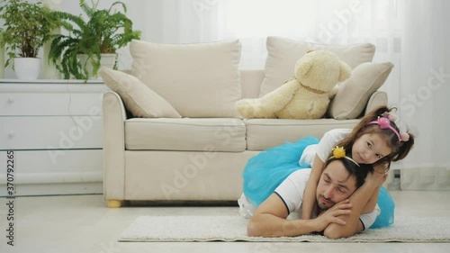 Pretty little girl with pigtails is lying on her father's back. Father is tired and wants to sleep, but daughter is full of energy and wants to play. photo