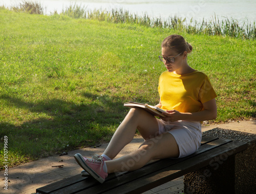 A beautiful young woman student in glasses, reading a book, sits on a bench, against the background of a summer green park on a sunny day in the shade of trees.